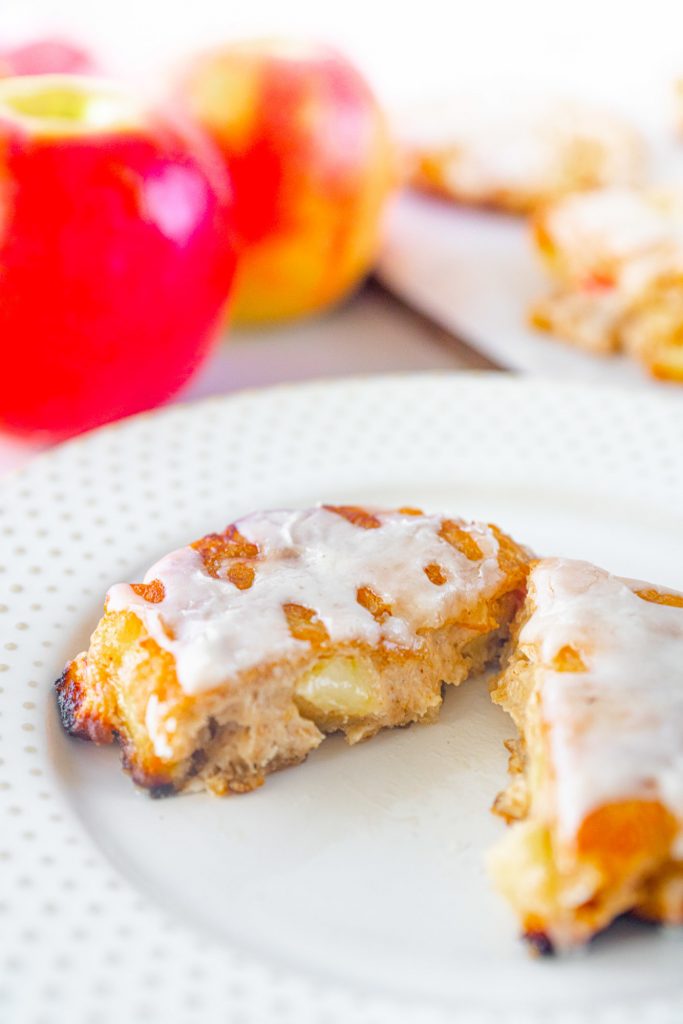 Single vegan apple fritter cut in half so you can see what the inside looks like on a white plate with red apples in the background.