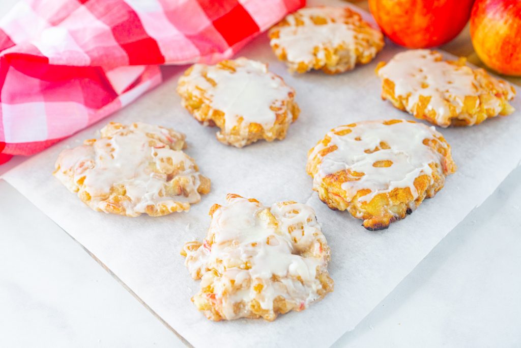 Six vegan apple fritters on cooling tray with apples and towel in the background.