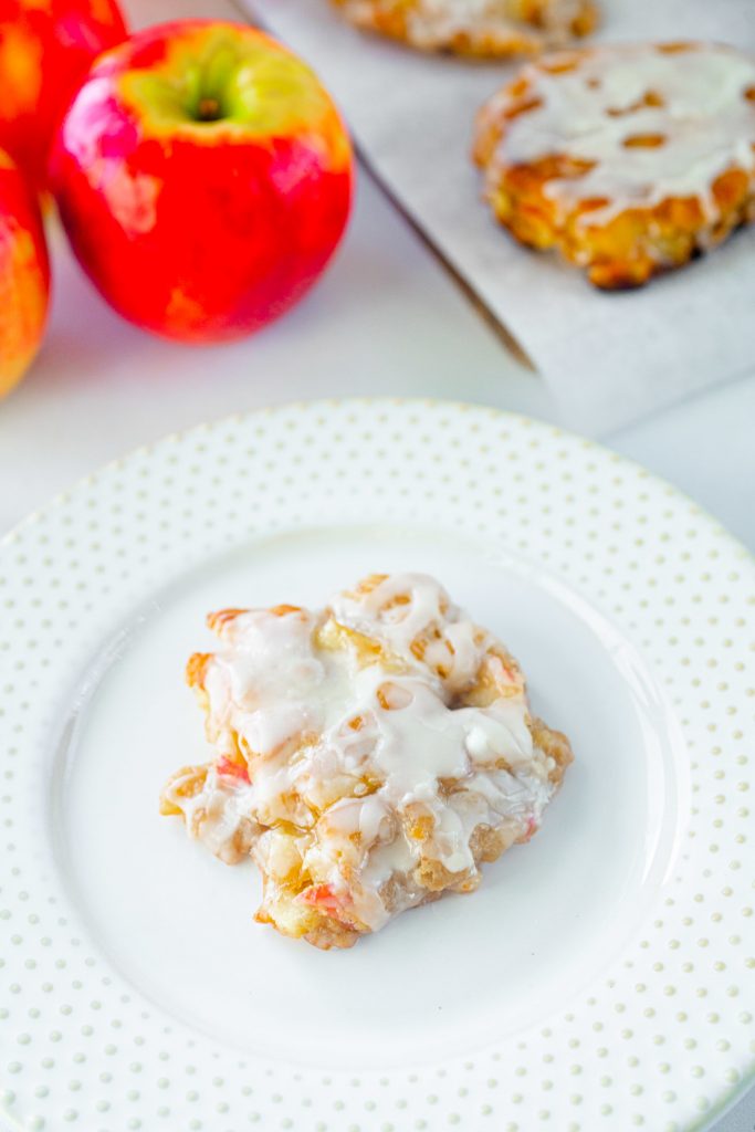 single vegan apple fritter on white plate with apples in the background