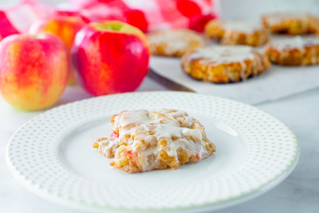 side view of a single apple fritter on a white plate with more in the background with apples.