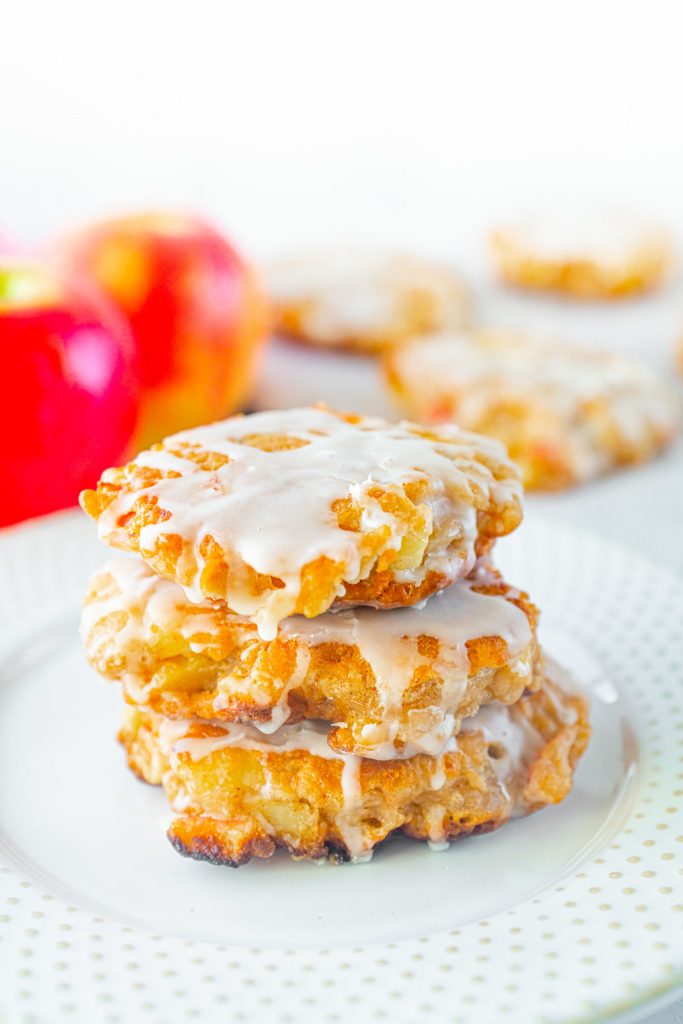 vegan apple fritters stacked on top of each other and glazed on a white plate with apples in the background.