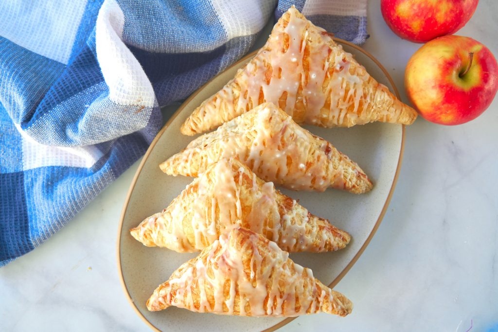 four glazed apple turnovers from above on tan plate with blue towel and red apple