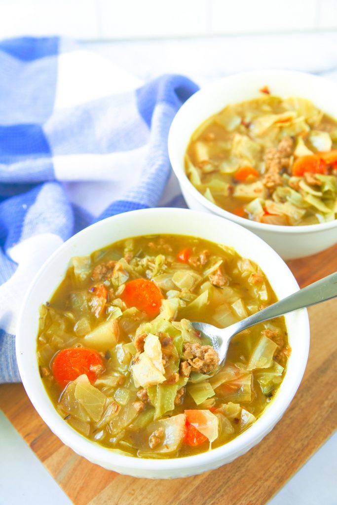 two bowls of vegan cabbage soup one with a spoon on cutting board next to a blue and white towel.