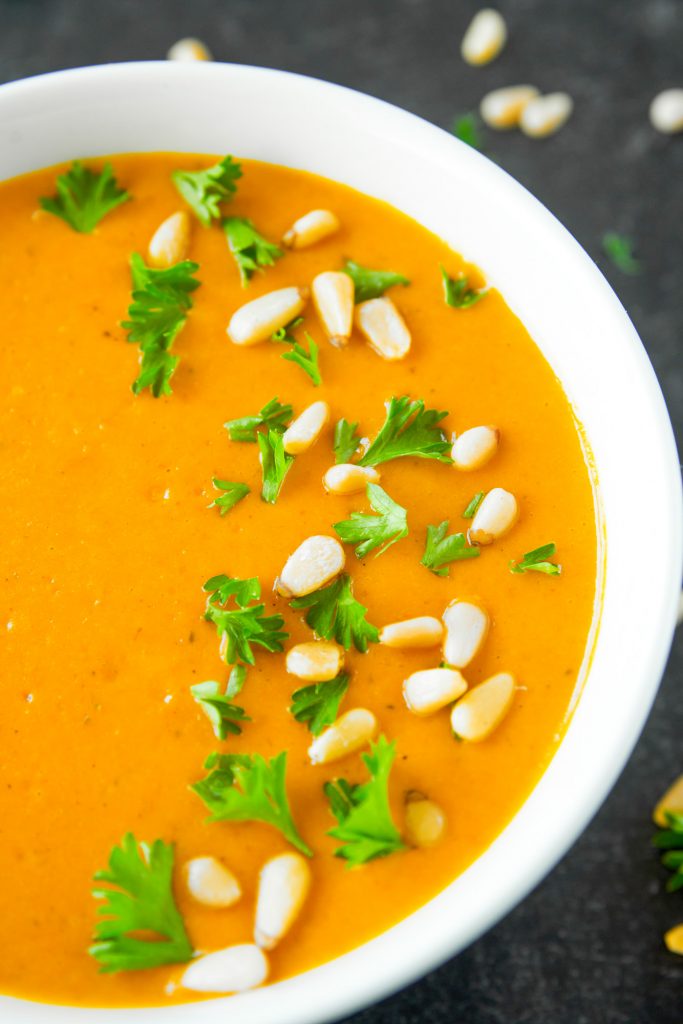 Close up of white bowl full of orange vegan carrot soup with pine nuts and parsley sprinkled on top.