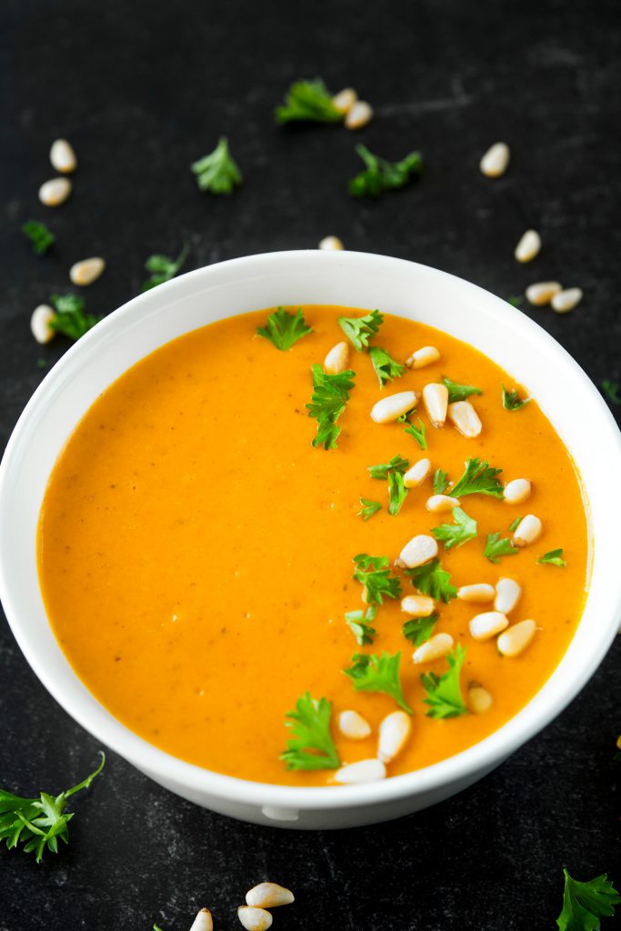 White bowl full of orange vegan carrot soup with pine nuts and parsley sprinkled on top.