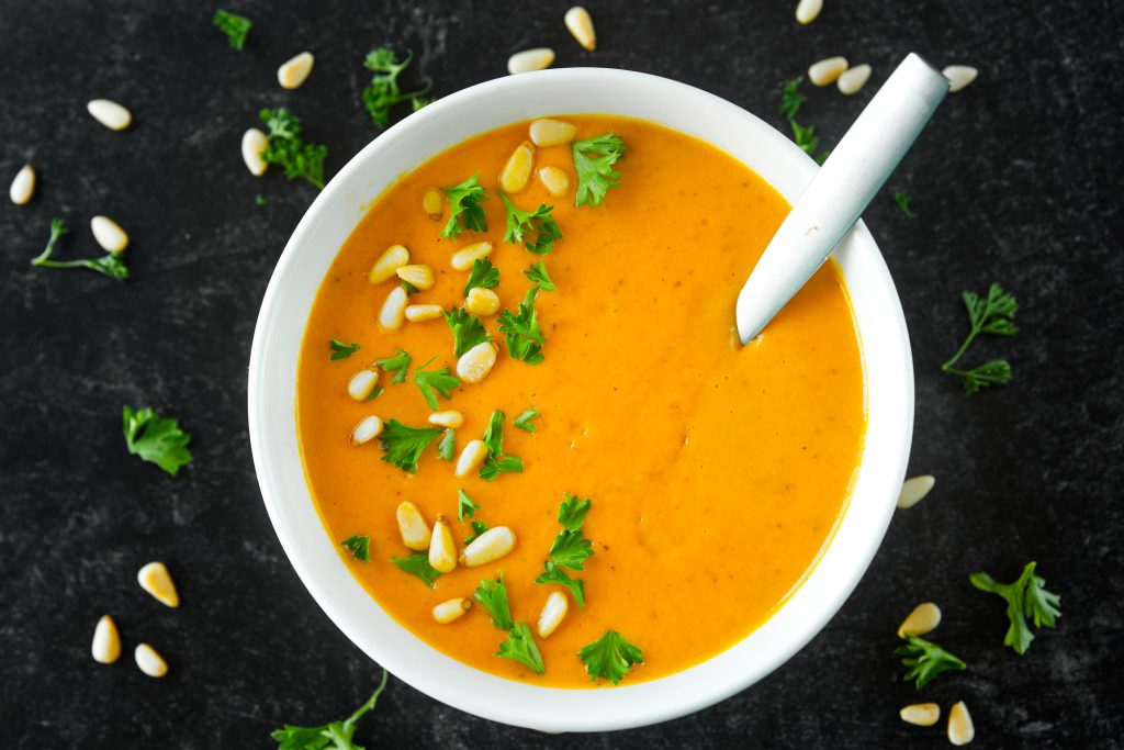 Straight down photo of white bowl full of orange vegan carrot soup with pine nuts and parsley sprinkled on top with a spoon.