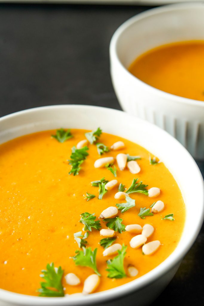 Close up ofhite bowl full of orange vegan carrot soup with pine nuts and parsley sprinkled on top.