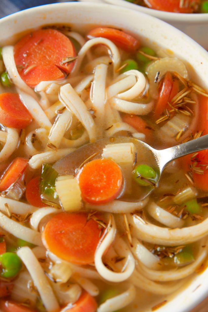 close up of spoon scooping up vegetables and noodles from a bowl of soup.