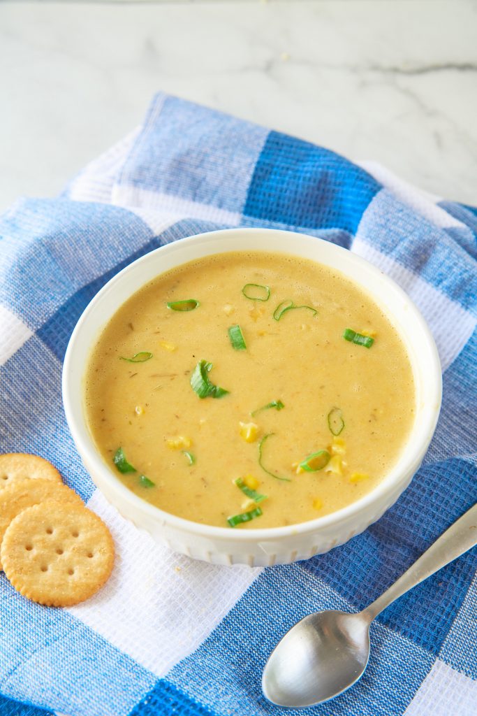 Vegan corn chowder in a white bowl with spoon and crackers on a plaid blue and white towel.