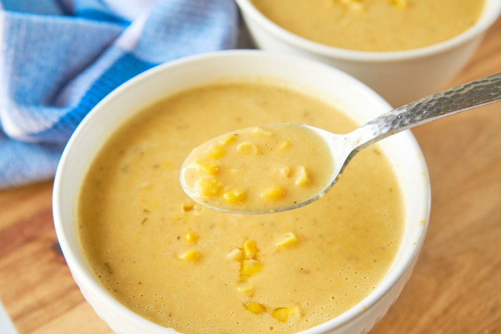 A spoon scooping up yellow vegan corn chowder from a white bowl.