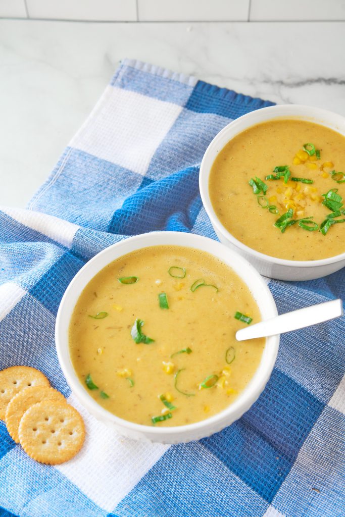 two white bowls of corn chowder with one spoon on white and blue towel with crackers.