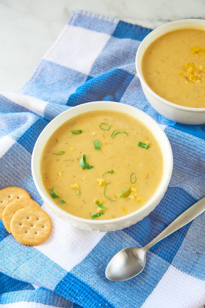 two white bowls of yellow corn soup on plaid white and blue towel with a spoon and crackers.