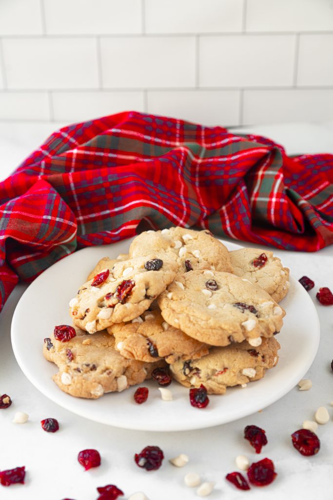 White plate of vegan white chocolate cranberry cookies next to a tartan towel with cranberries and white chocolate chips scattered around.