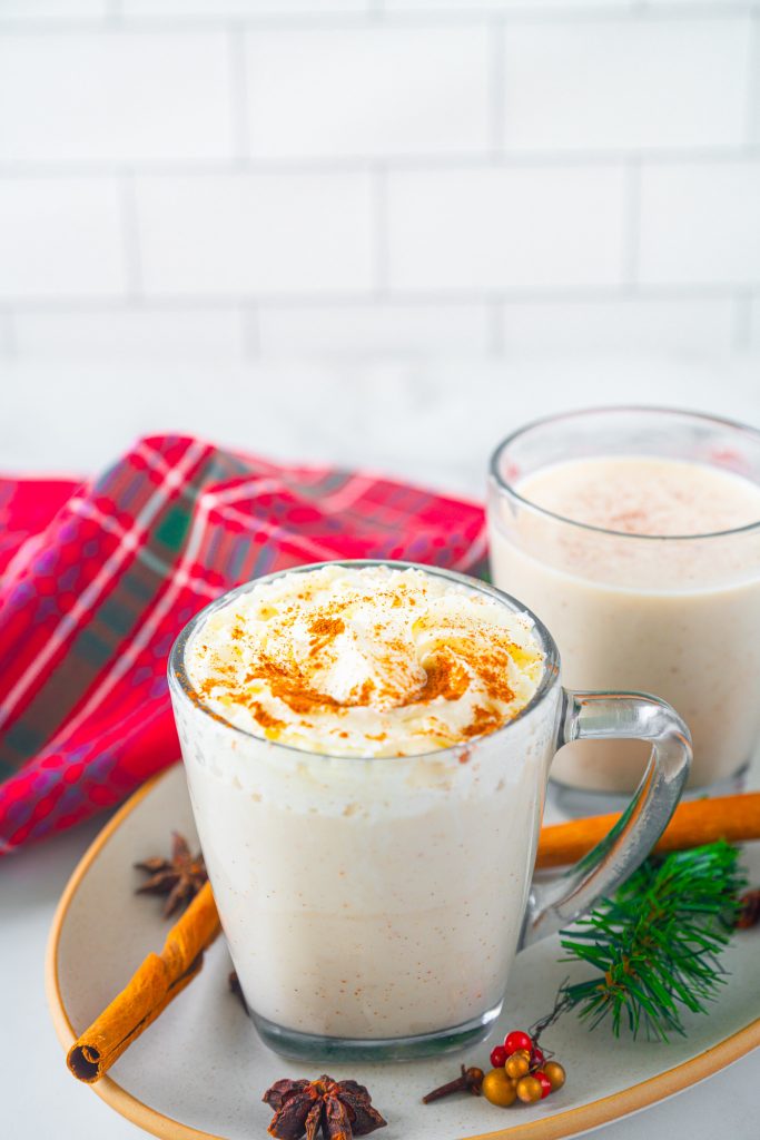 two cups of vegan eggnog on a serving tray one with whipped cream and cinnamon and one without