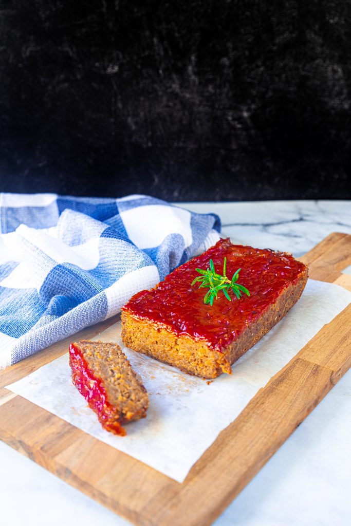 sliced vegan meatloaf recipe with black background. the nut loaf is sitting on a brown cutting board with blue dish towel