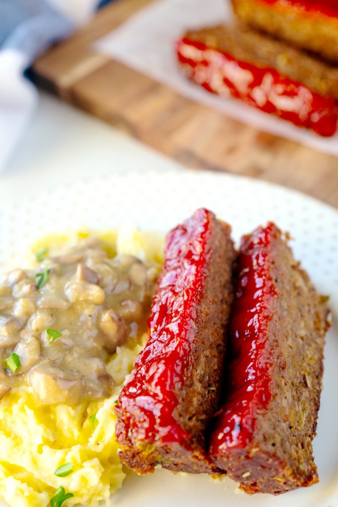 close up of vegan meatloaf on mashed potatoes with meatless meatloaf sliced in the background with red glaze