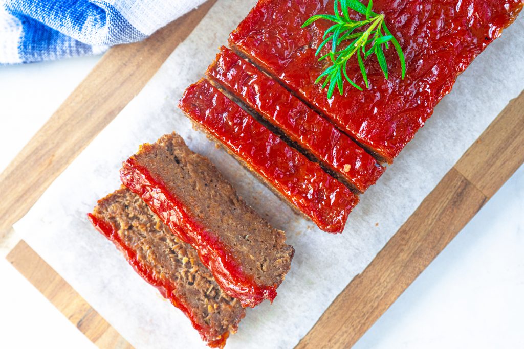 red glazed meatless meatloaf from above with slices and rosemary on a brown cutting board with a blue dish towel