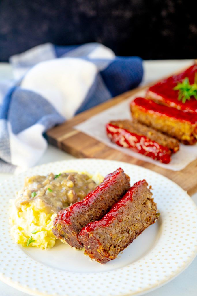 vegan meatloaf slices on a bed of mashed potatoes with veggie loaf in the background with blue towel on counter