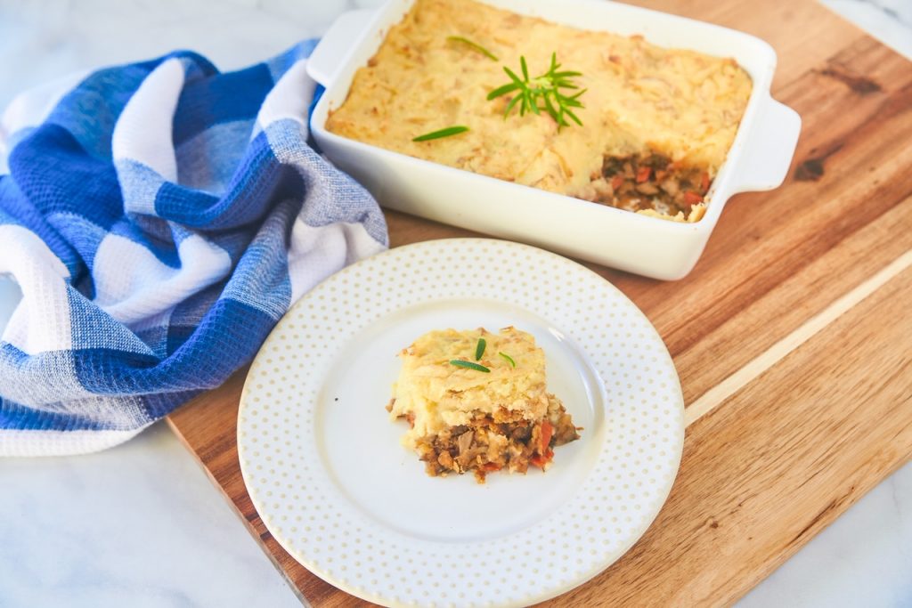 A slice of vegan cottage pie on white plate with casserole and blue hand towel in the background.