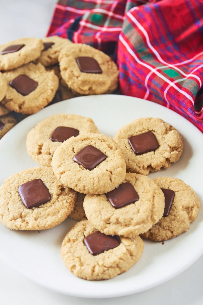 Vegan peanut butter blossoms on white plate with other cookies in the background with a tartan cloth. The cookies are round and brown with a square of chocolate in the middle.