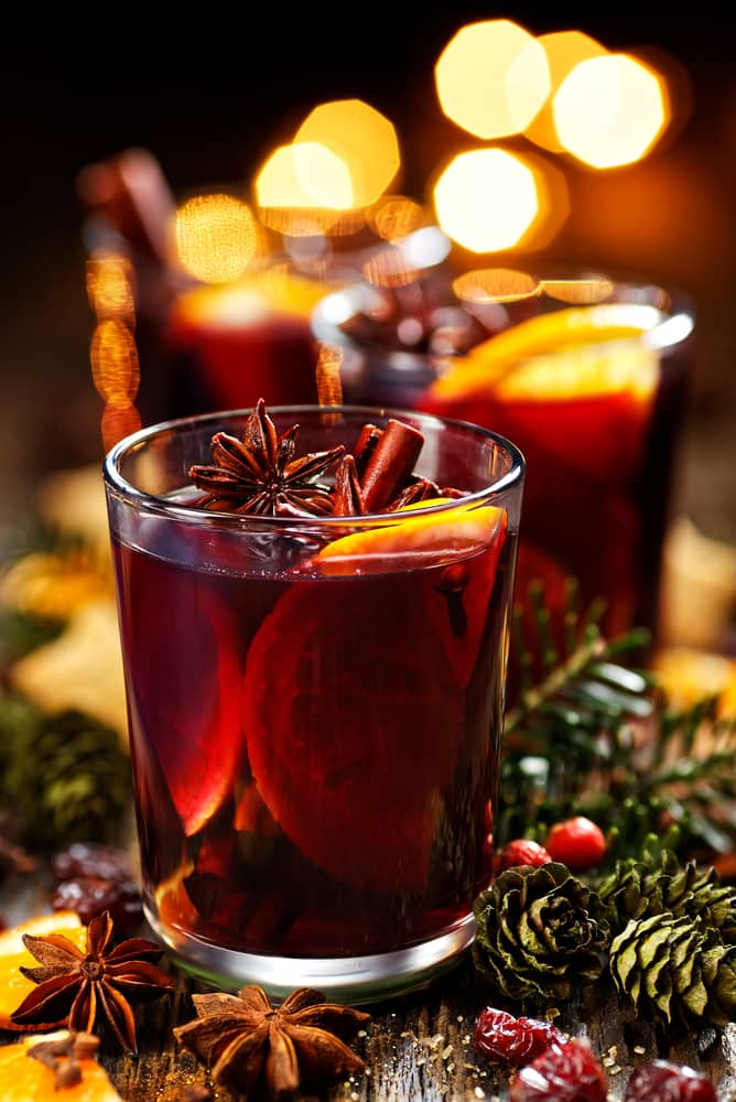 Close up of mulled wine in a glass with fruit and cinnamon garnish with Christmas lights in the background.