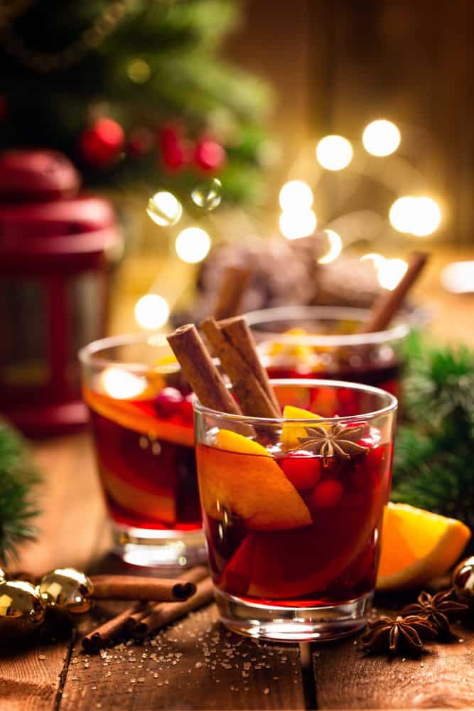 Three glasses of Gluhwein mulled wine garnished with cinnamon and full of fruit with Christmas lights in the background.