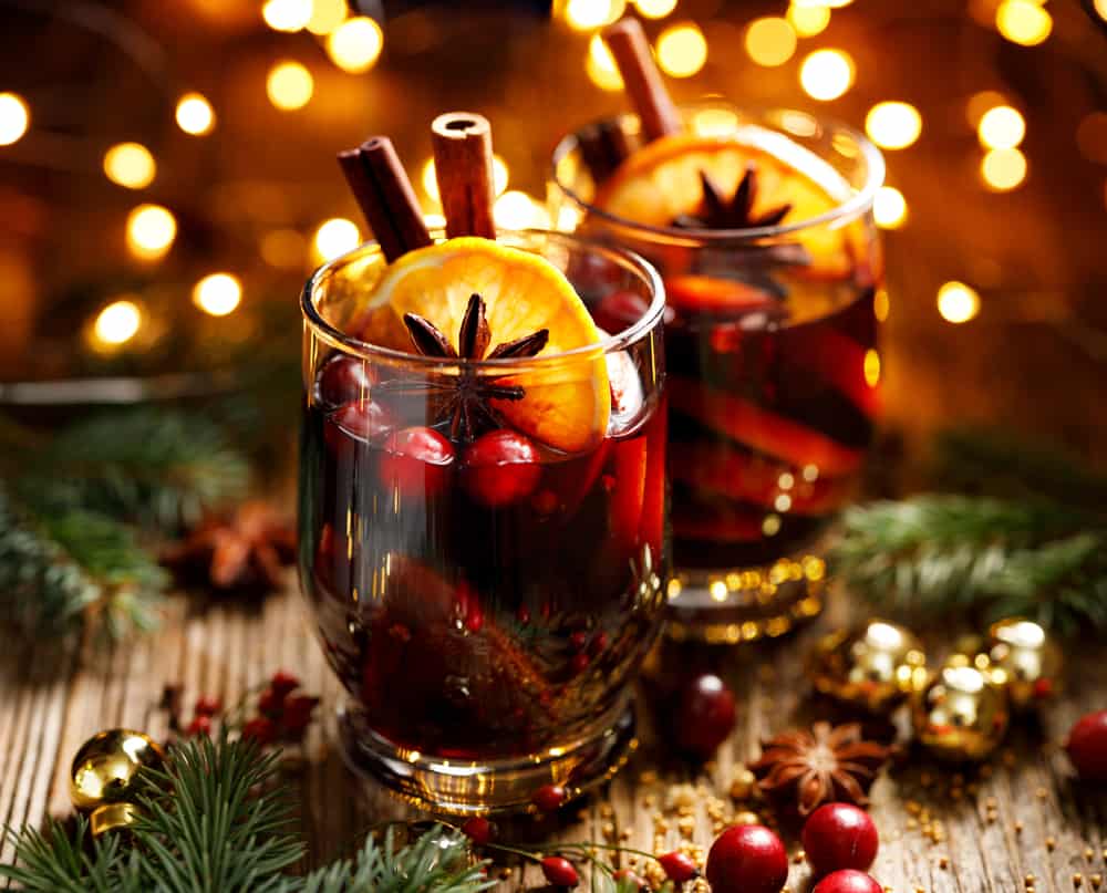 Two short glasses of Gluhwein with oranges, cranberries, and cinnamon sticks with pieces of fir branches and Christmas lights in the background.
