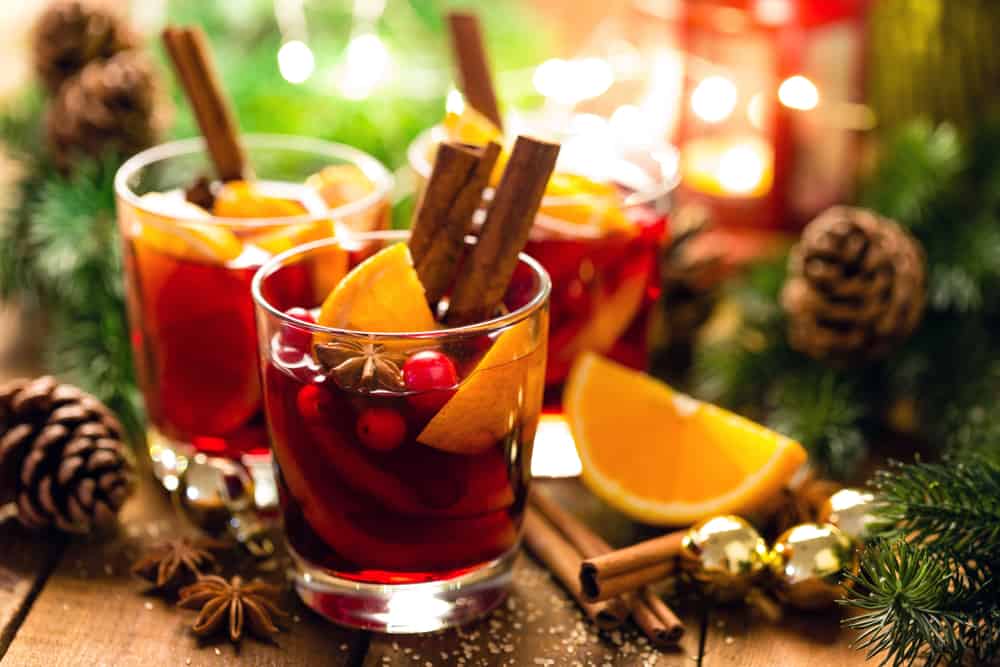 Three glass cups of Gluhwein on a table with fir branches, pine cones, cinnamon sticks, orange slices, and golden bells.