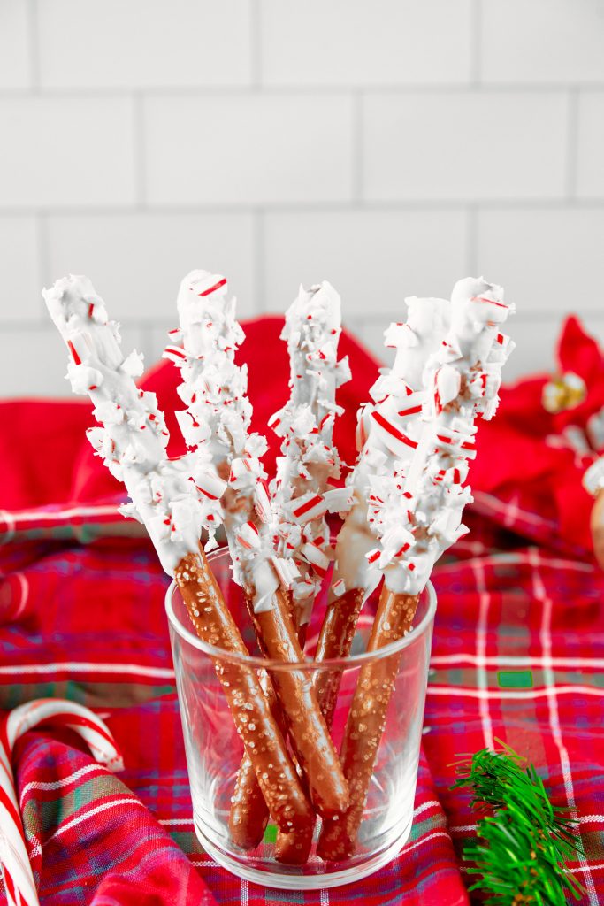 Christmas pretzel rods coated in white chocolate and covered with crushed peppermint in a glass jar on a tartan towel.