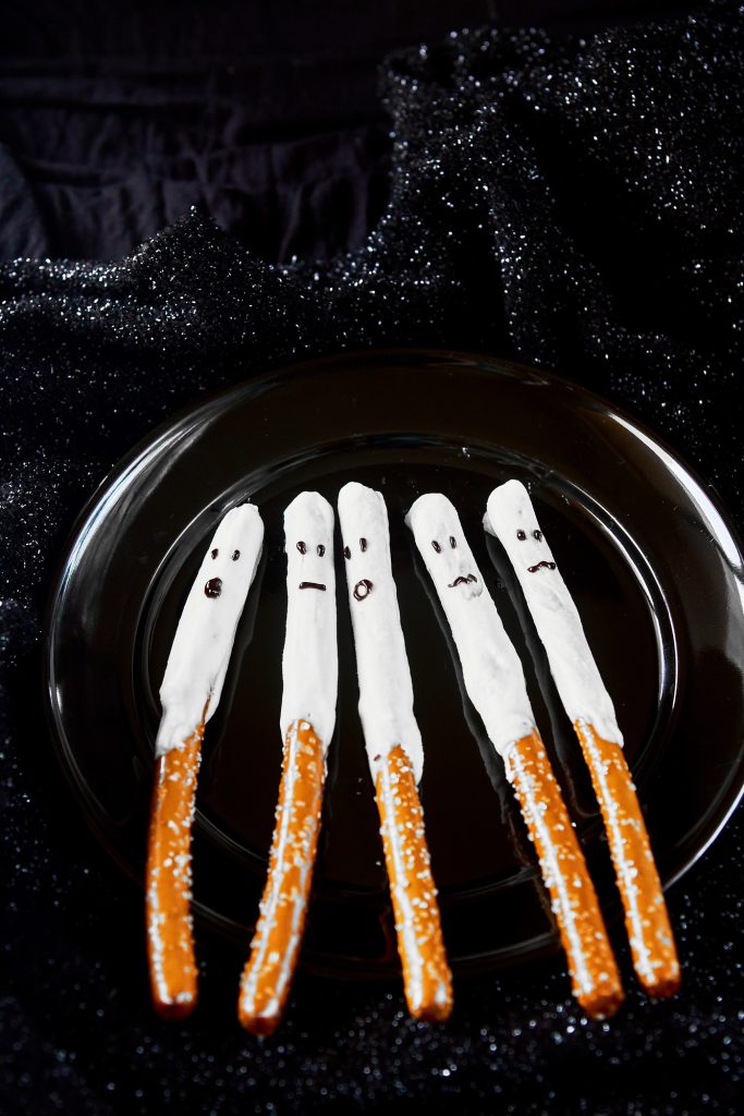 cute halloween pretzels that look like ghosts dipped in white chocolate on a black plate with sparkly counter behind