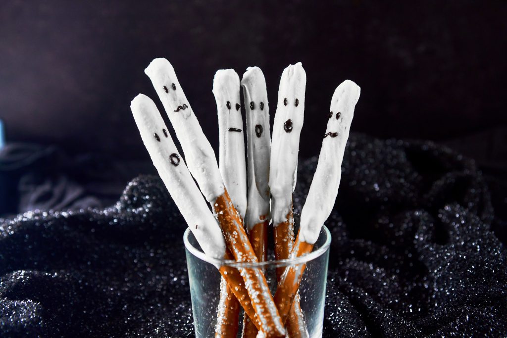 six halloween pretzel rods dipped in white chocolate so they look like ghosts in a glass on a black background 