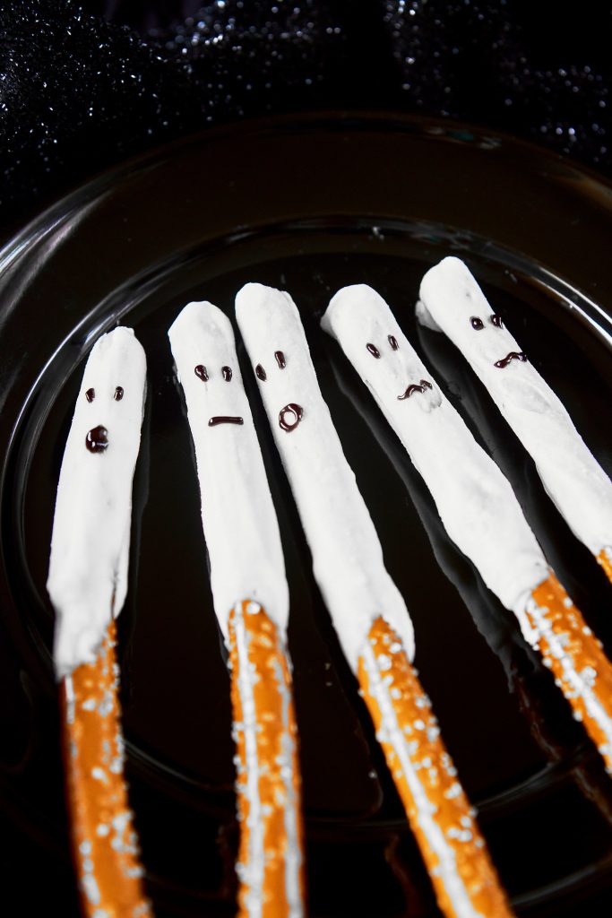 five ghost halloween pretzels covered with white chocolate with little faces drawn on sitting on a black plate