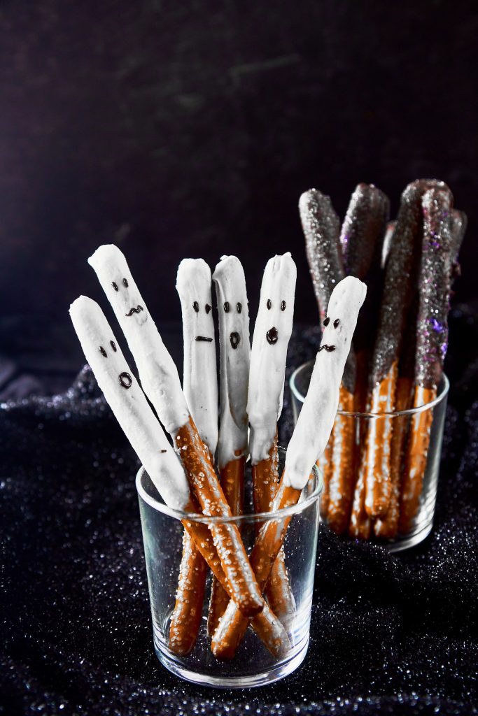white chocolate dipped pretzel rods that look like ghosts standing up in a glass with dark chocolate halloween pretzel rods in the background