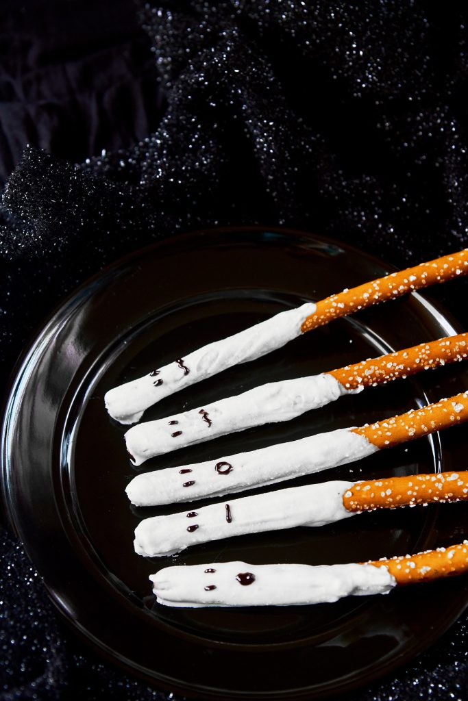 cute white chocolate dipped halloween pretzel sticks with faces drawn on to look like ghosts on a black plate 