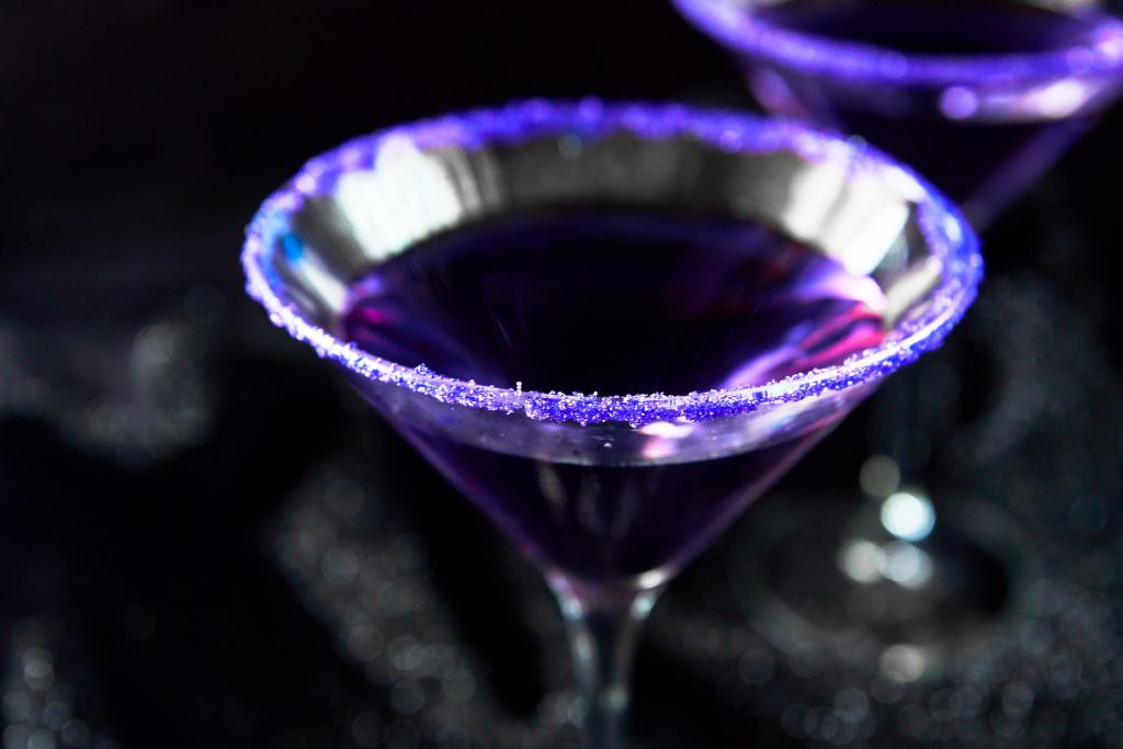 A close up of Purple people eater Halloween cocktail in glass with purple sugar on rim against a black background.