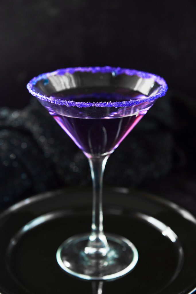 Purple people eater Halloween cocktail in glass with purple sugar on rim against a black background.