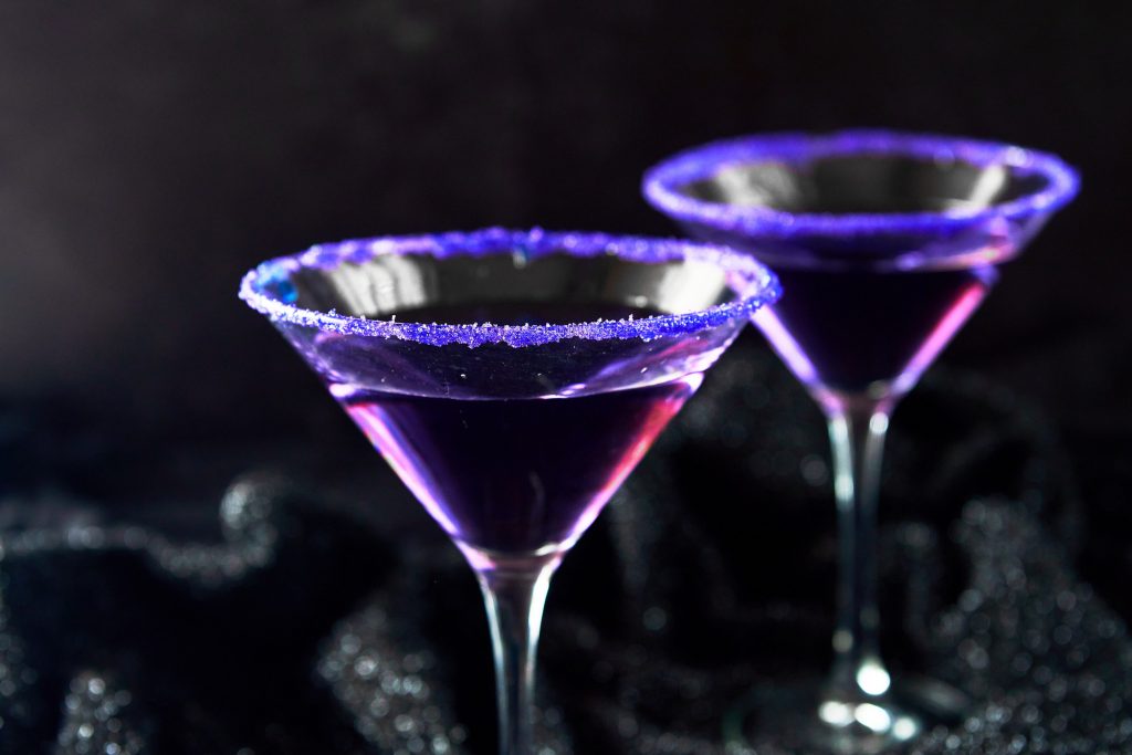 Two purple people eater cocktails in glass with purple sugar on rim against a black background.