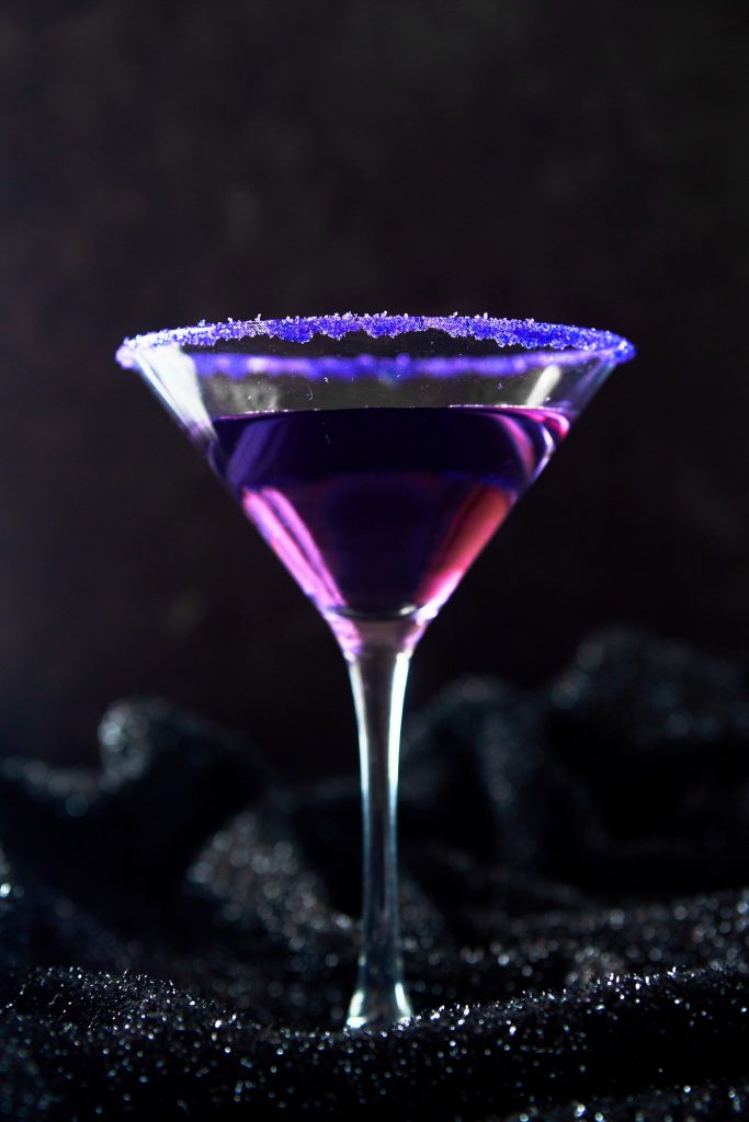 Purple cocktail in glass with purple salt on rim against a black background.