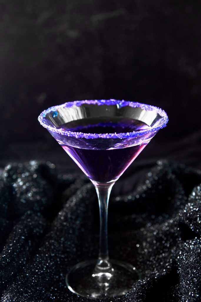 striking Purple people eater Halloween cocktail in glass with purple sugar on rim against a black background.