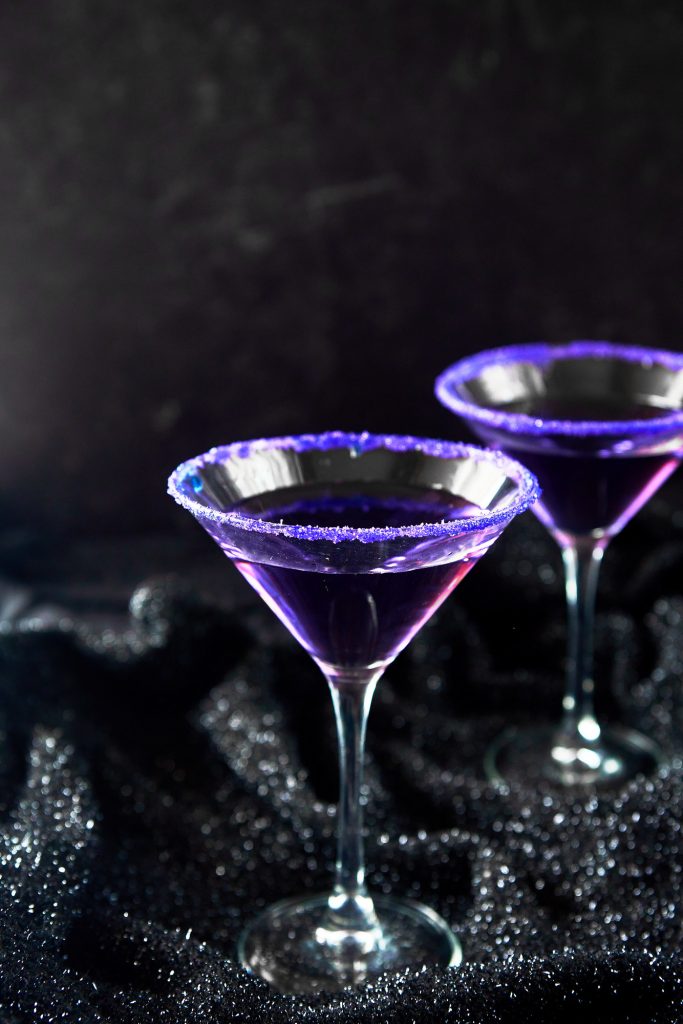 Two Purple people eater Halloween cocktails in glasses with bright purple sanding sugar on rim against a black background.