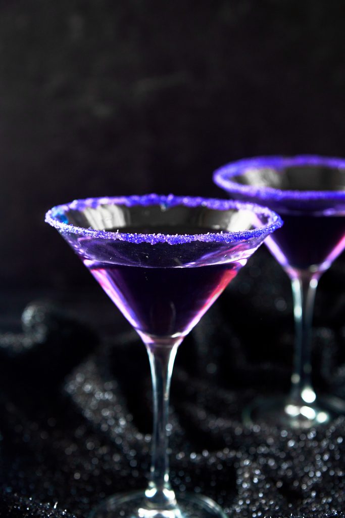 two purple people eater Halloween cocktail in glass with purple sugar on rim against a black background.