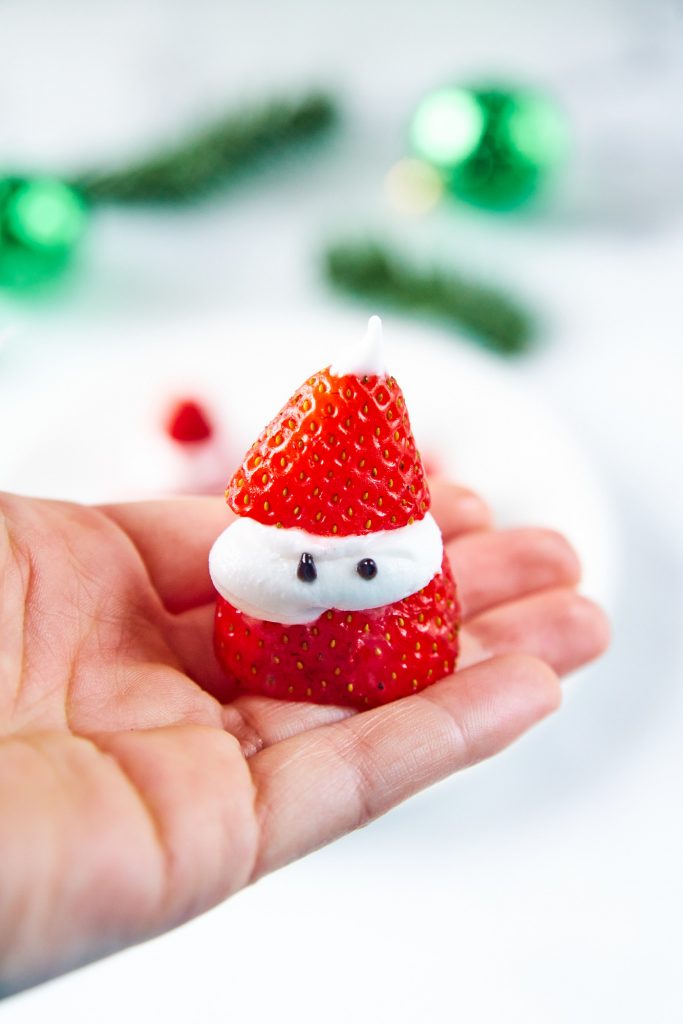hand holding a strawberry that looks like Santa Claus with white cream and black icing eyes