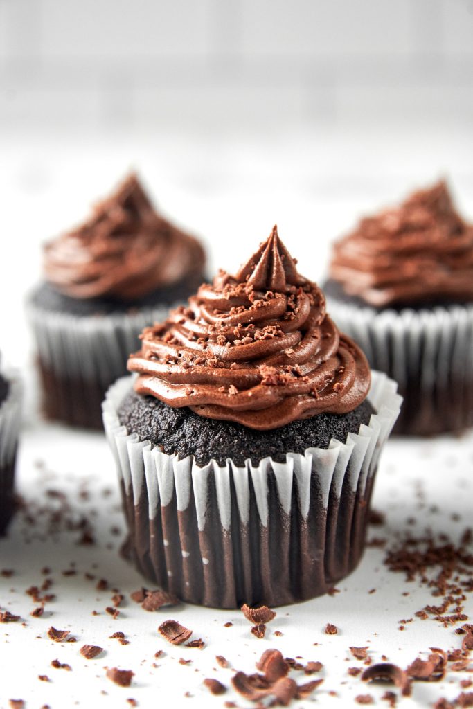 A vegan chocolate cupcake piled with frosting and more cupcakes in the background with chocolate shavings all around.