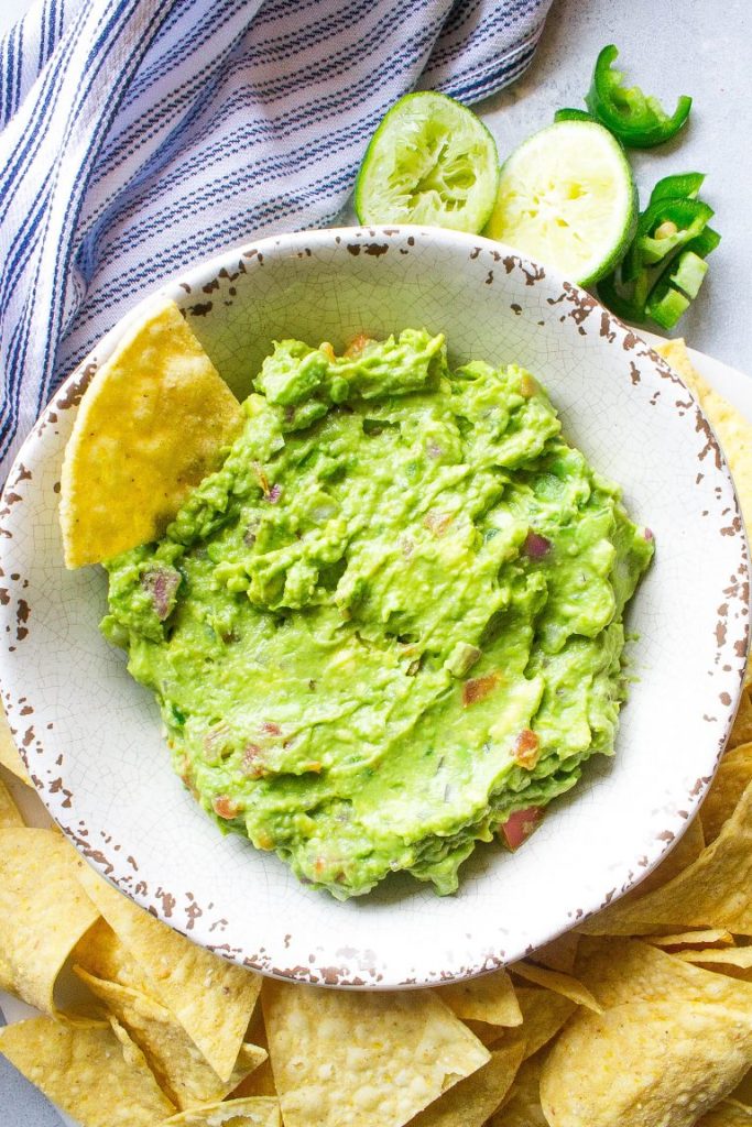Photo of guacamole being served in a white bowl surrounded by tortilla chips.