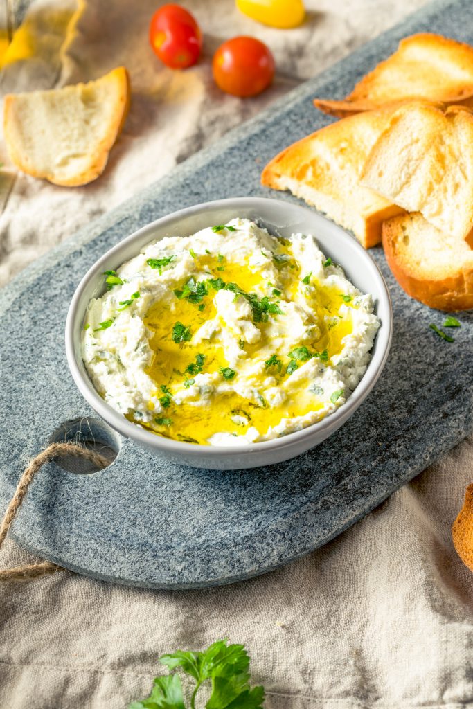 Grey bowl holding goat cheese dip with olive oil drizzle on plate with bread.