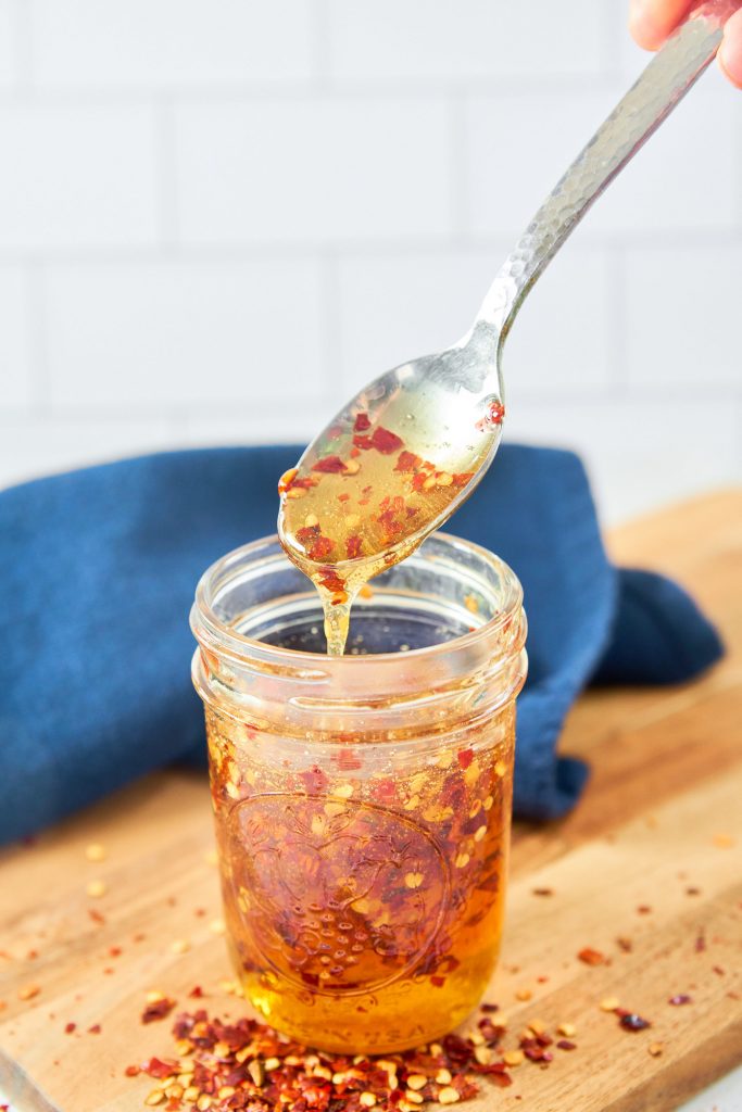 A spoon scooping up hot honey from a jar with red pepper flakes on a cutting board.