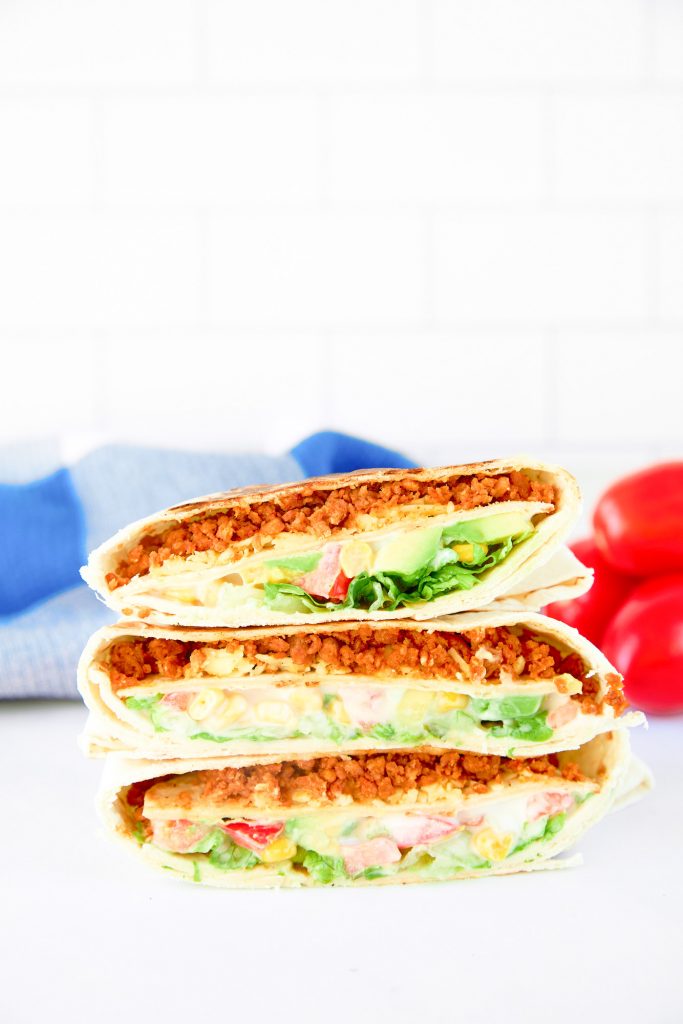 vegan crunchwrap supreme stacked on a white counter with blue towel and tomatoes