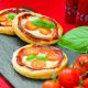 Photo of Mini Margherita Puff Pizza Pies. 3 mini margherita pizzas are seen on a slate board with a red table cloth underneath. Vine tomatoes and a bright green basil leaf is on the right of the image.