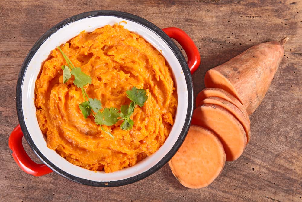 mashed sweet potatoes in a red bowl with chopped sweet potato on the outside of a brown counter top