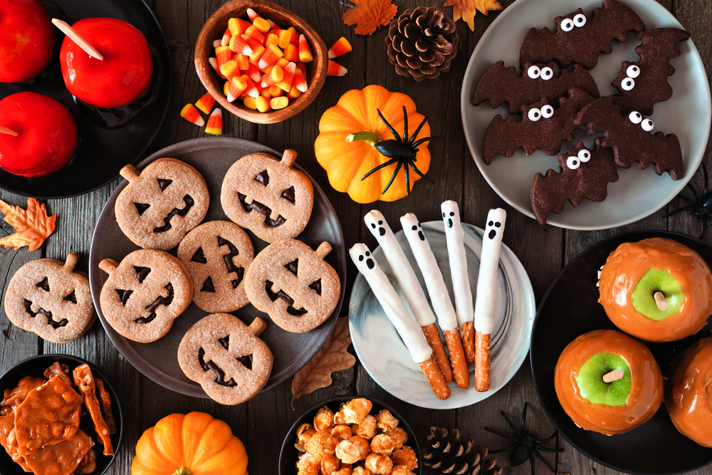 five white chocolate dipped halloween pretzel rods on a white plate surrounded by a variety of other halloween snacks and candied apples 